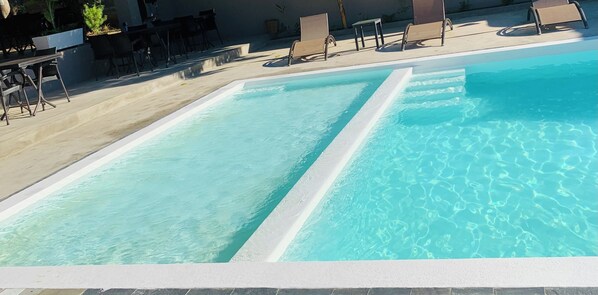 Una piscina rectangular con agua cristalina y azul, rodeada de sillas de descanso y una tumbona.