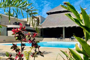 Un área de piscina con una estructura de techo de paja, palmeras y un edificio moderno al fondo.