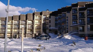 A multi-story residential building with balconies, a parking area with a car, and a snowy ground.