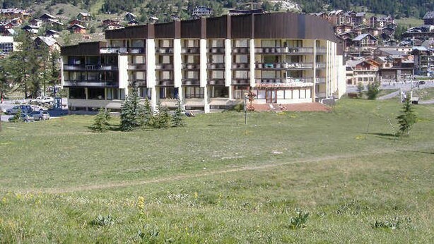 A multi-story hotel with a flat roof and balconies, surrounded by a grassy field and a mountainous backdrop.