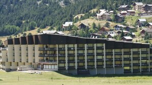 A large hotel with multiple floors and balconies, surrounded by a green landscape and a forested hill in the background.