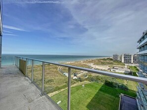 Varanda com vista para a praia e oceano, área gramada e edifícios modernos ao fundo.