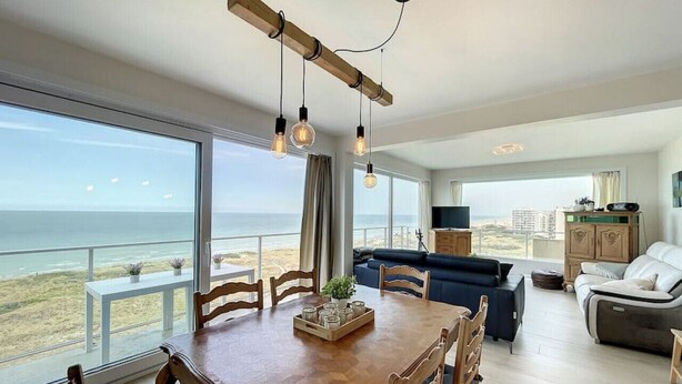 A spacious living area with a dining table, chairs, and a sofa. There is a view of the ocean through large windows. - Flat Sunset - amazing seaview (De Panne)