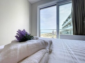 Quarto de hotel com cama, janela com vista para a praia e edifícios, e uma planta na cama.