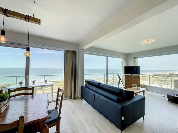 A spacious living room with a large window overlooking the beach, a dark sofa, a wooden dining table with chairs, and a flat-screen TV on a wooden cabinet. - Flat Sunset - amazing seaview (De Panne)