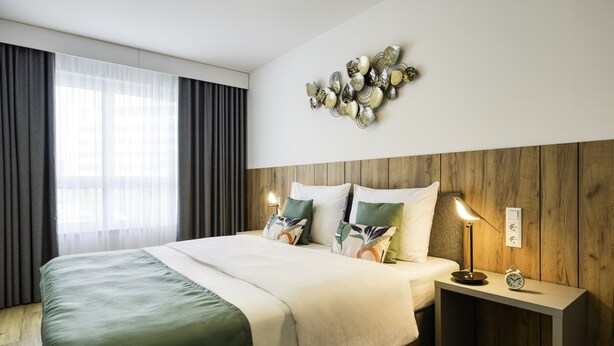 A hotel room with a wooden headboard, a large bed with a green and white bedspread, a bedside table with a lamp, and a wall-mounted clock. - Somerset Schoenbrunn Vienna (Vienna)