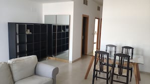A modern living room with a sofa, a glass dining table with chairs, a black shelving unit, and a door leading to another room.