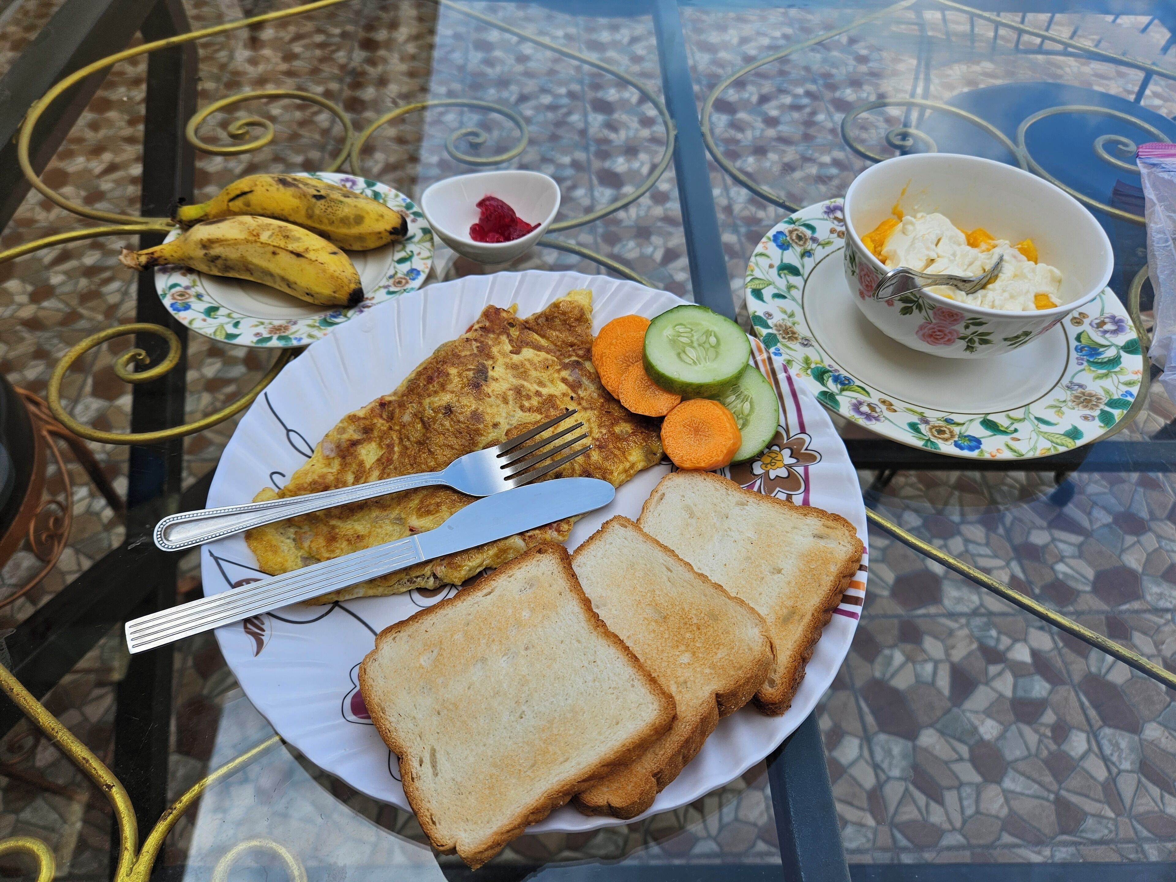A breakfast plate with toast, a fried egg, sliced cucumbers, carrots, and bananas on a table with a patterned tablecloth.