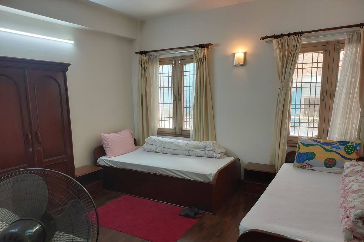 A bedroom with two beds, a wooden wardrobe, a standing fan, and a window with curtains.