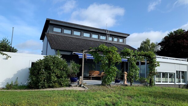 Ein modernes, zweistöckiges Haus mit geneigtem Dach, großen Fenstern und einem gepflegten Garten mit Topfpflanzen und einer Bank.