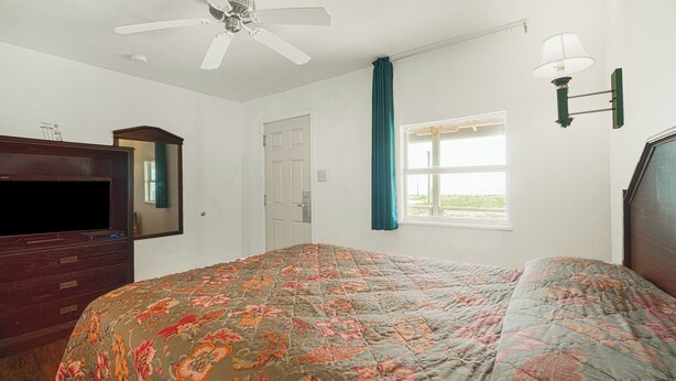 Quarto com cama, ventoinha no teto, penteadeira, televisão, janela com cortinas e luminária na parede.