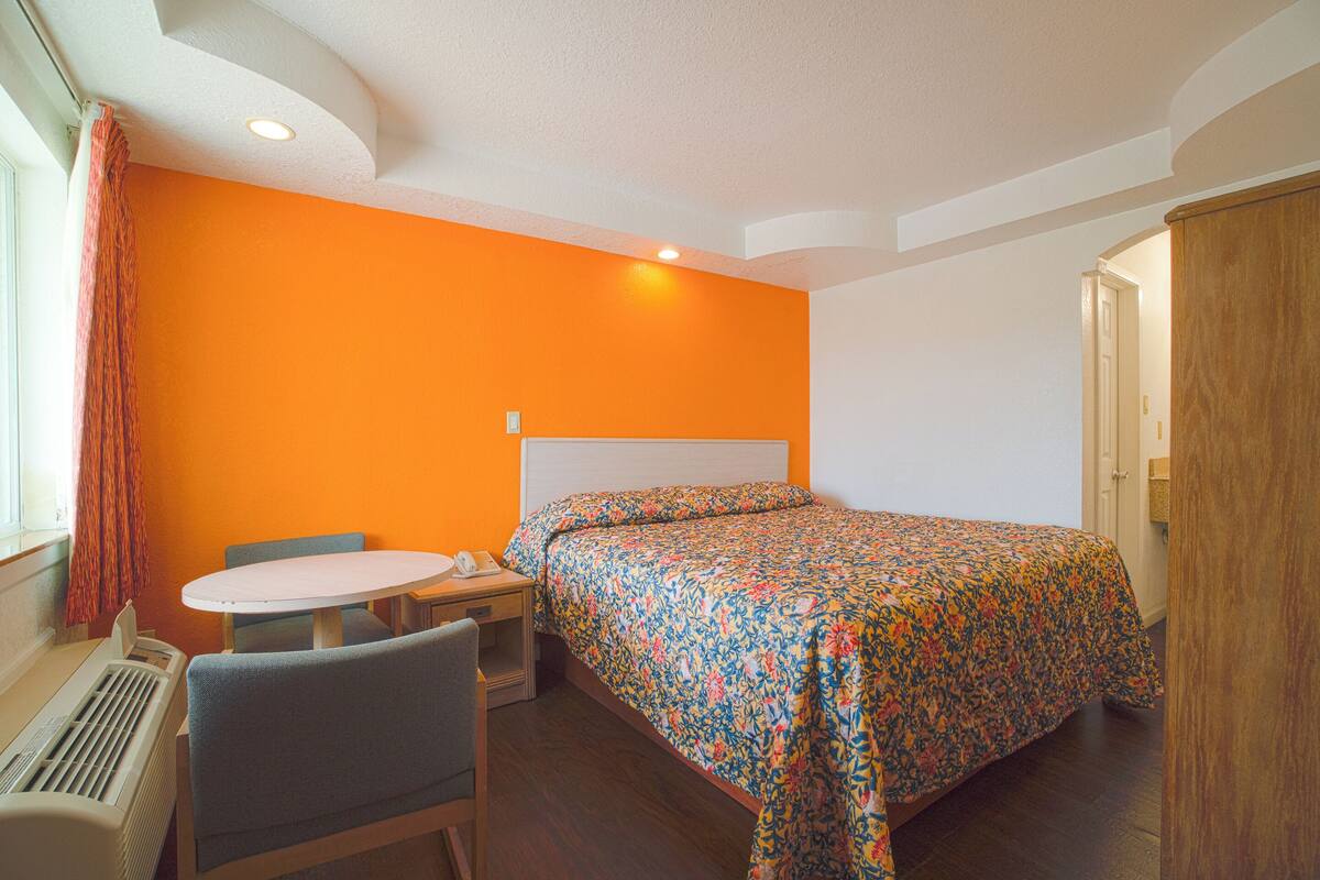 A hotel room with a bed, a desk with a chair, a window with curtains, and a radiator.
