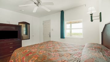Un dormitorio con cama, ventilador de techo, cómoda, televisor, ventana con cortinas y una lámpara de pared.