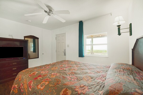 A bedroom with a bed, a ceiling fan, a dresser, a TV, a window with curtains, and a wall-mounted lamp.