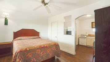 A bedroom with a wooden bed, a ceiling fan, a nightstand, a lamp, and a kitchenette with a stove and cabinets.