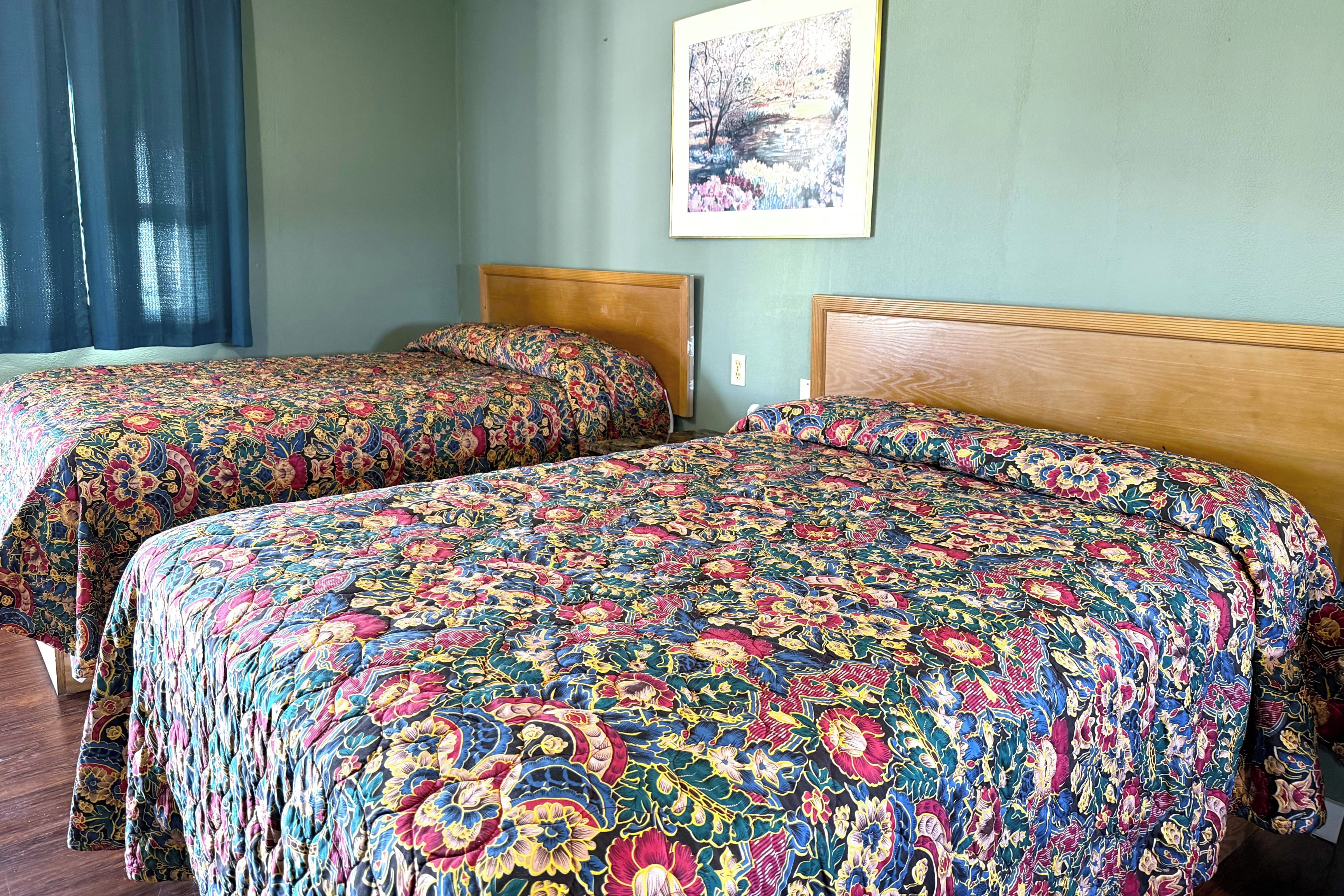 Two double beds with floral-patterned bedding, a wooden headboard, and a framed picture on the wall.