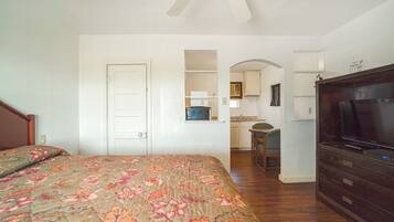 A bedroom with a bed, a dresser, a television, and a kitchen area visible through an archway.