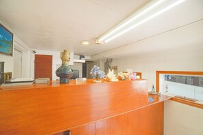 A reception desk with a wooden finish, a vase, and a small plant. There is a door in the background and a window with a view. - Budget Inn By Oyo Corpus Christi Beach (Portland)
