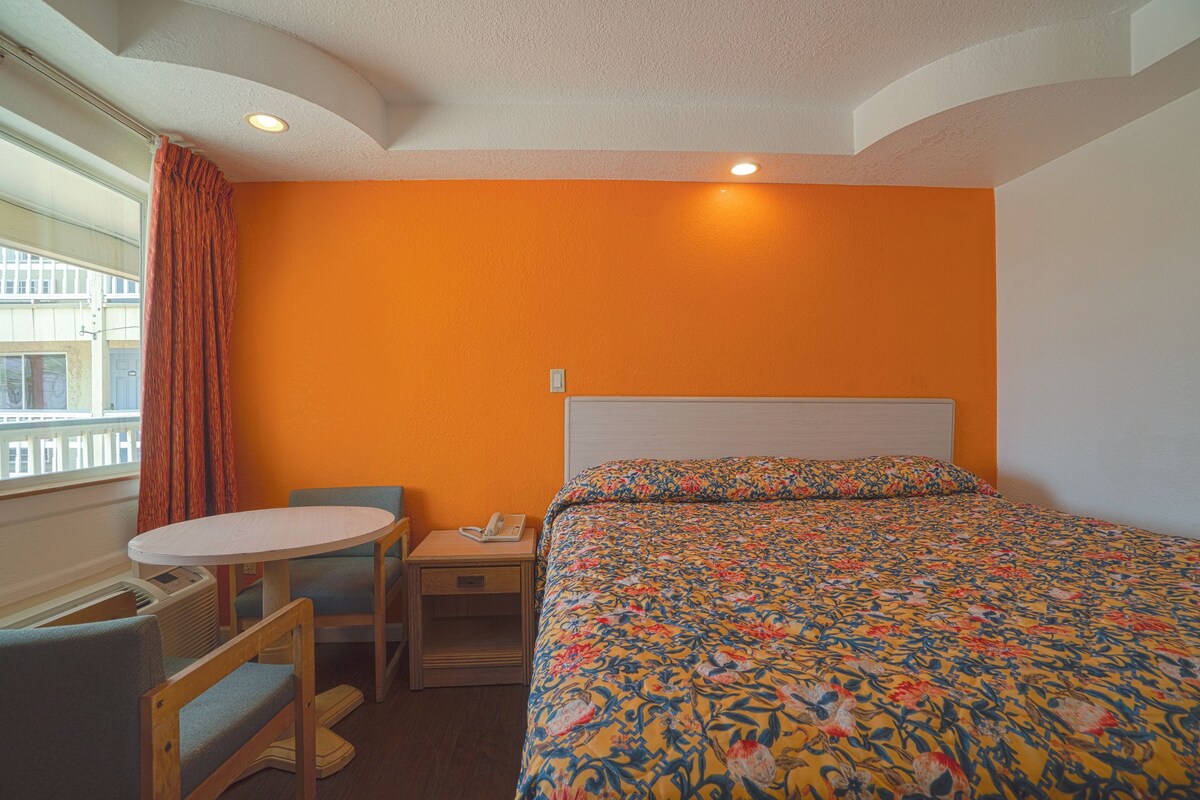 A hotel room with an orange wall, a bed with a floral bedspread, a small table with a chair, and a window with a curtain.