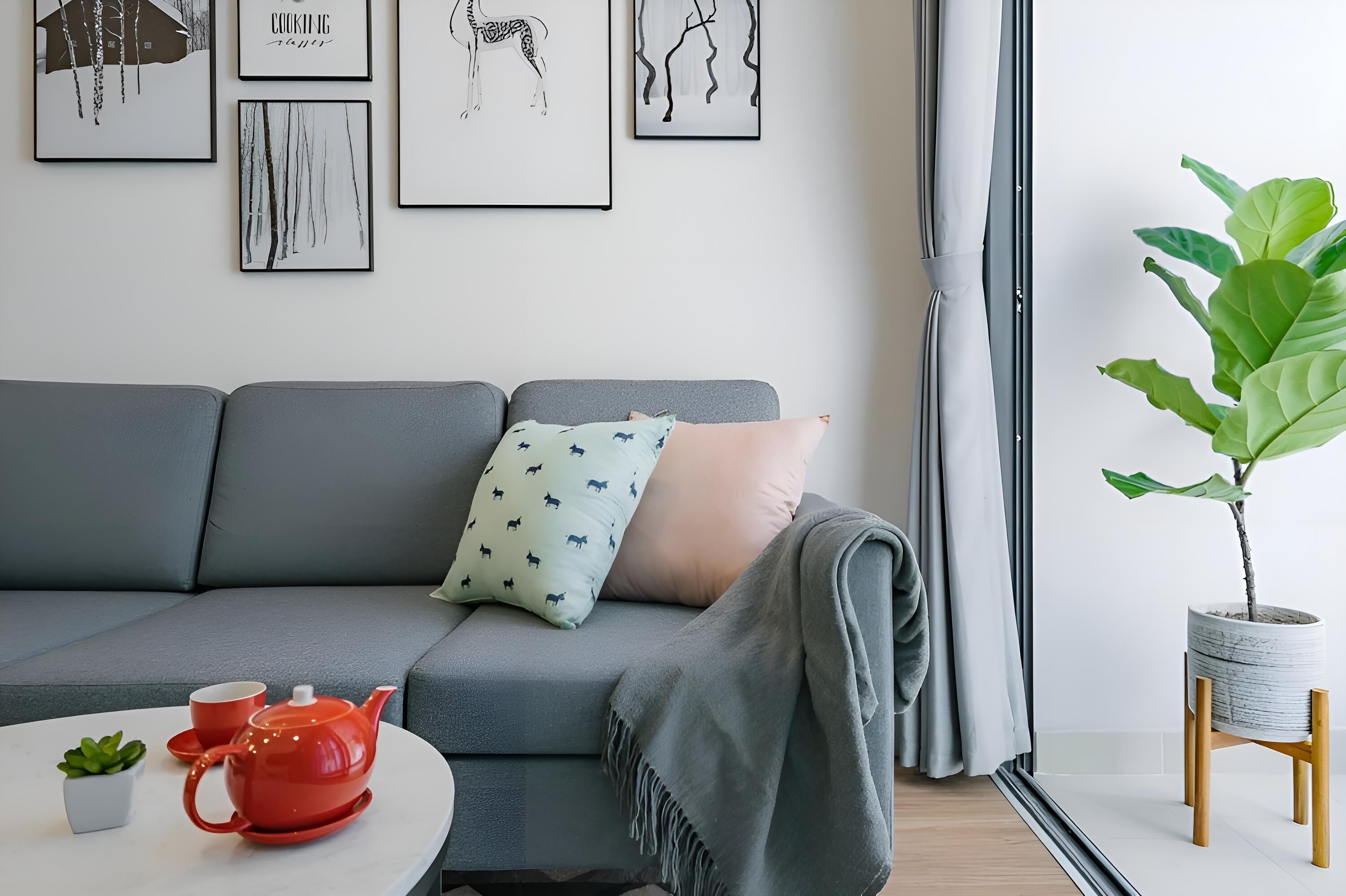 Un salon moderne avec un canapé gris, une petite table ronde sur laquelle se trouvent une théière et une tasse rouges, et une plante en pot.