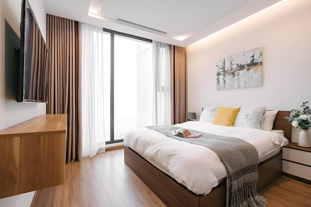 A modern bedroom with a large bed, a TV mounted on the wall, and a sliding glass door leading to a balcony.