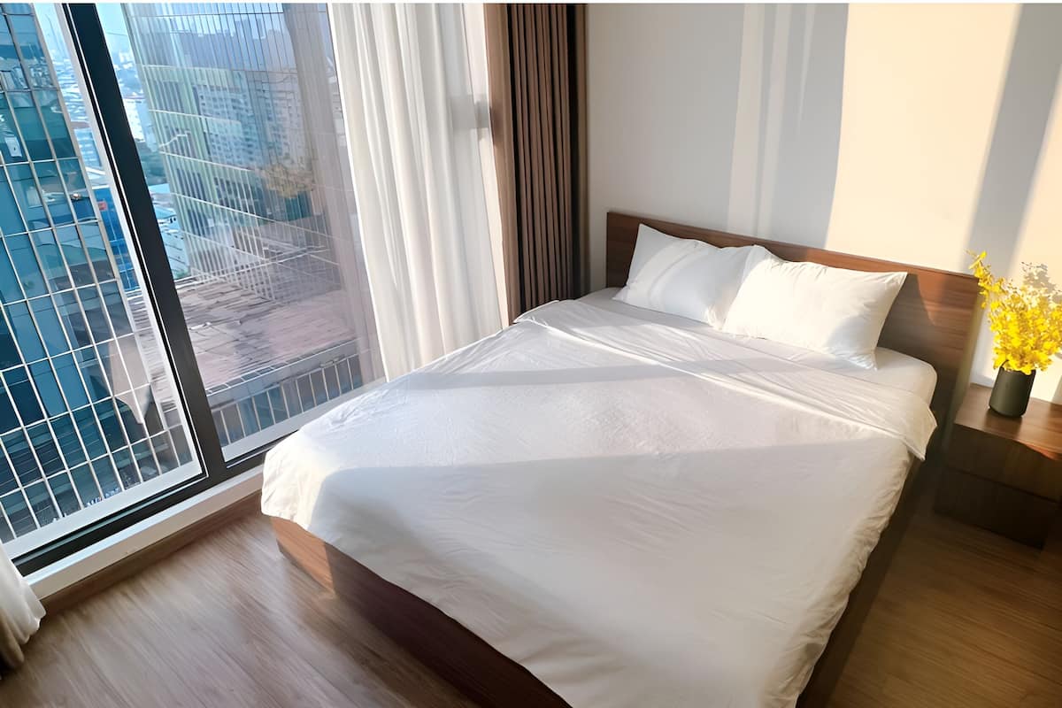 A hotel room with a large bed, wooden headboard, white bedding, a nightstand with a vase of yellow flowers, and a view of a cityscape through a large window with curtains.