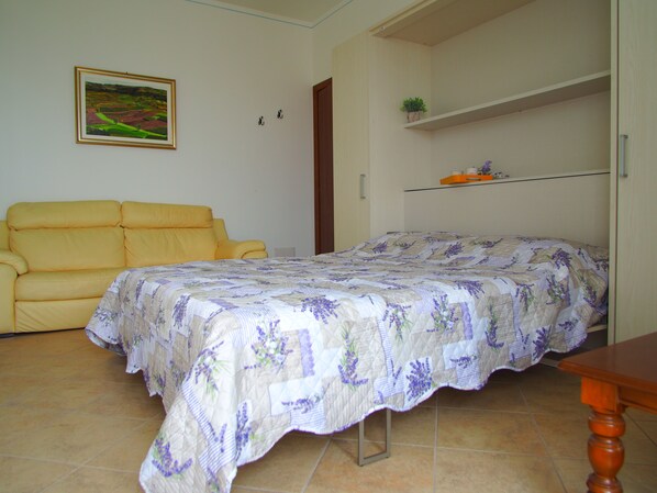 A bedroom with a bed, a yellow sofa, a painting on the wall, and a shelf with a plant. - Parson's Vineyard Brown B (Verduno)
