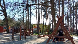 A playground with swings and climbing structures, surrounded by trees and a yellow building in the background.