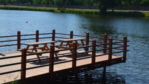 A wooden dock with picnic tables extending over a calm lake, surrounded by lush greenery and a fence.