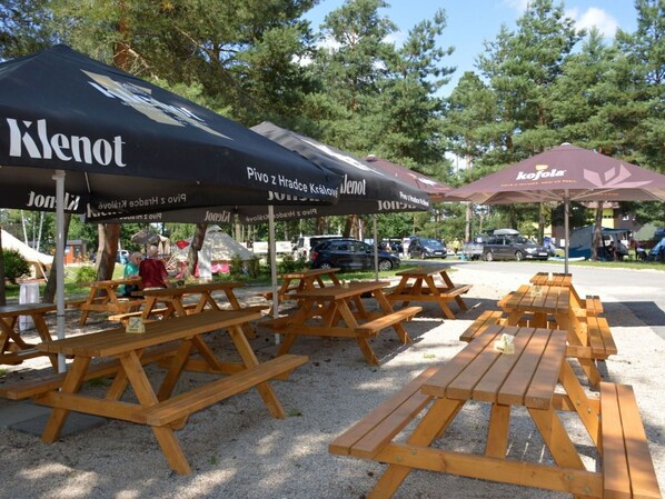 Terrassen tafels en parasols met logo's, gelegen in een parkachtig gebied met bomen en geparkeerde auto's op de achtergrond.