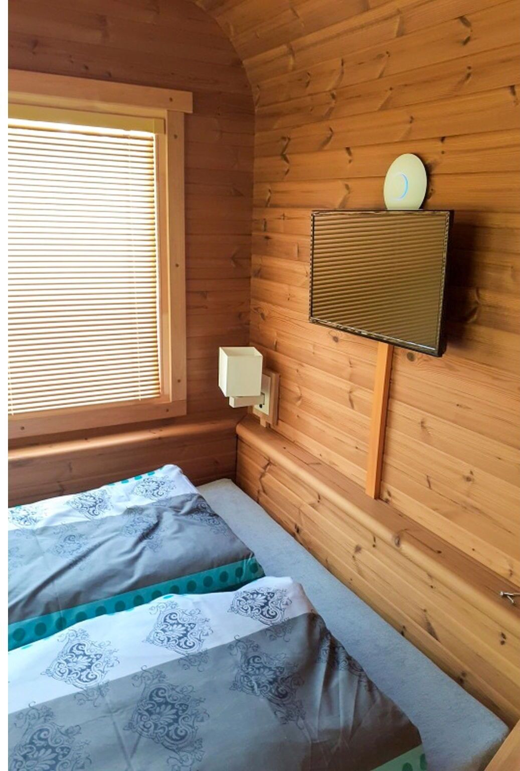 A wooden cabin room with a bed, a window with blinds, and a wall-mounted speaker.