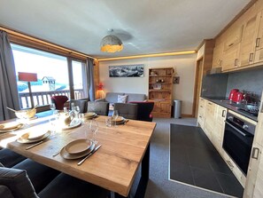Cozinha moderna com armários de madeira, sala de jantar com mesa de madeira posta para quatro pessoas e vista do exterior através de grandes janelas.