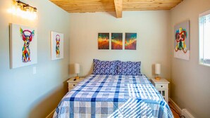 A bedroom with a wooden ceiling, a bed with a checkered bedspread, two wall-mounted lamps, and three framed artworks.