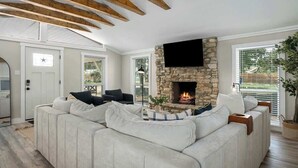 A modern living room with a stone fireplace, a large sectional sofa, and wooden beams on the ceiling. - Hummingbird Haus: Sleeps 12 with Hot Tub (Fredericksburg)