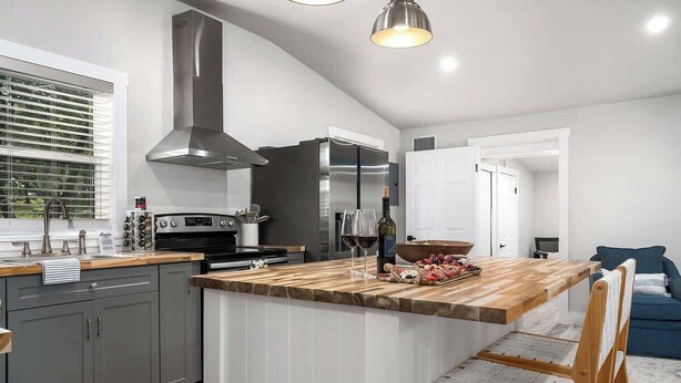A modern kitchen with grey cabinets, a stainless steel refrigerator, and a wooden island with seating. - Hummingbird Haus: Sleeps 12 with Hot Tub (Fredericksburg)