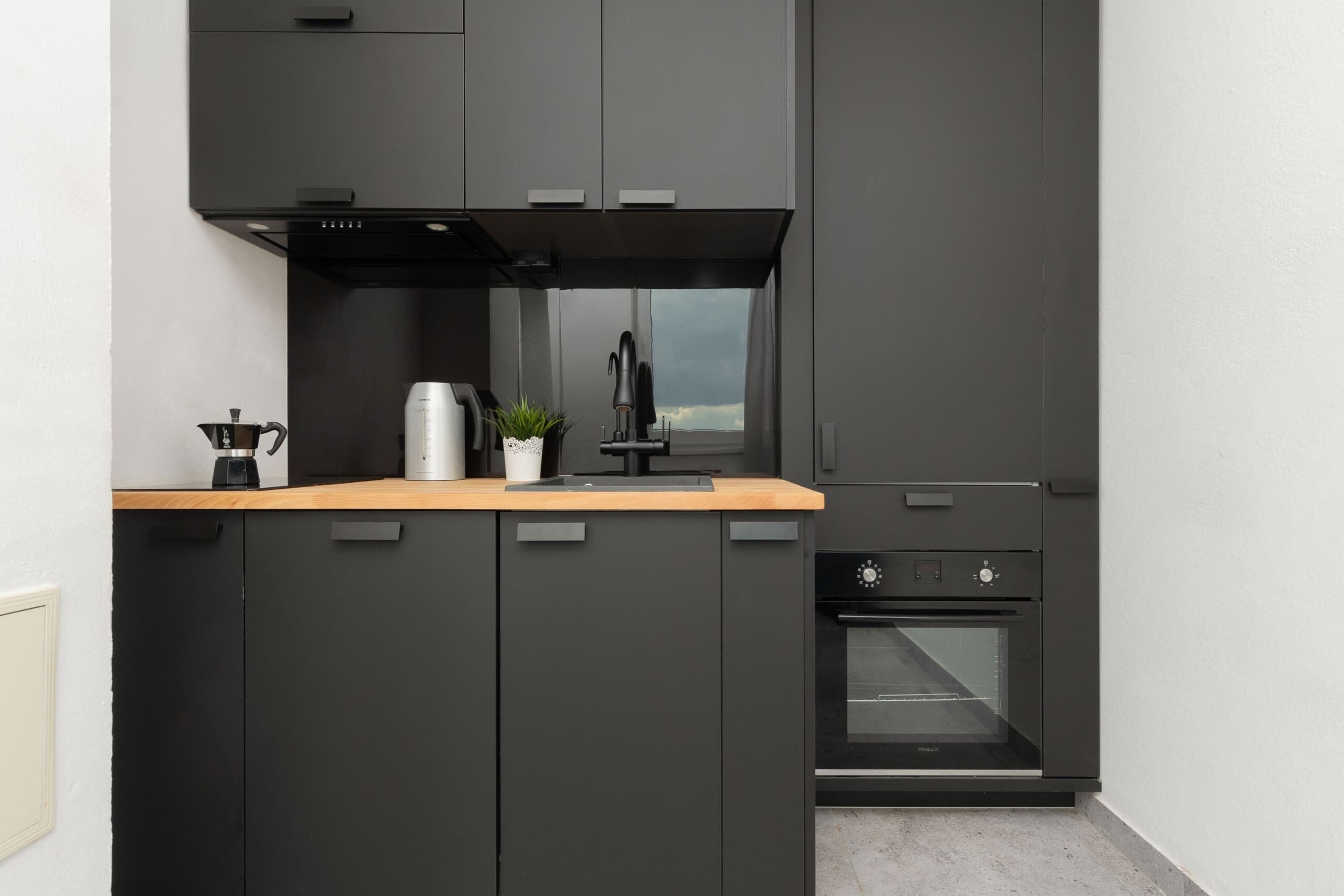 Une cuisine moderne avec des meubles de couleur sombre, un plan de travail en bois, une machine à café, une bouilloire, une petite plante en pot et un four encastré.
