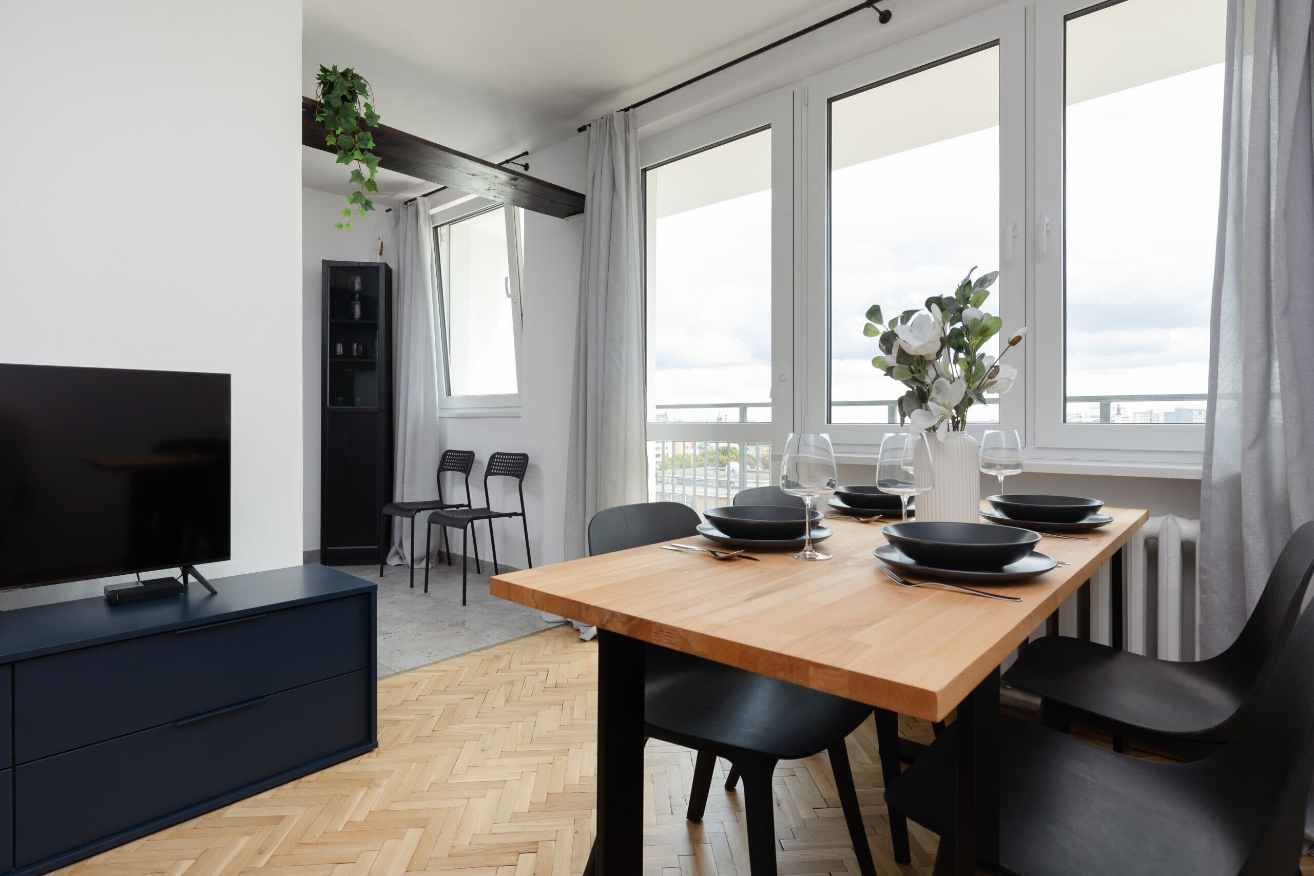 Un espace repas moderne avec une table en bois, des chaises noires et un téléviseur posé sur un meuble sombre.