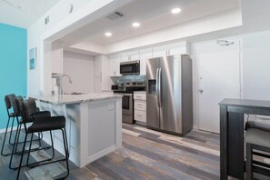 A modern kitchen with stainless steel appliances, a central island with bar stools, and a view of the adjacent living area. - Oceanfront Updated Condo - Direct Beach Access! (Satellite Beach)