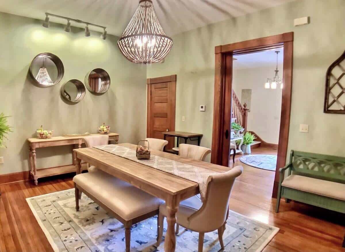 A dining area with a wooden table, benches, and a chandelier. There are two side tables with baskets and a mirror on the wall.