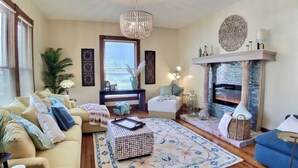 Un salon chaleureux avec un canapé, un fauteuil bleu, un tapis à motifs, une cheminée et un lustre suspendu.