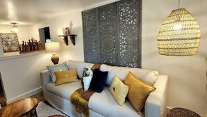 Un salon avec un canapé de couleur claire, des coussins décoratifs, un luminaire tressé et une étagère murale sur laquelle sont disposés divers objets.