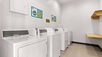 A laundry room with four white front-loading washers and dryers, a wooden shelf, and three signs on the wall.