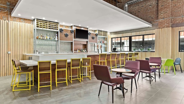 A modern bar area with a long counter, multiple seating options including yellow and purple chairs, and a brick wall backdrop.