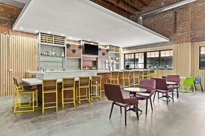 A modern bar area with a long counter, multiple seating options including yellow and purple chairs, and a brick wall backdrop. - Home2 Suites by Hilton Phoenix Downtown (Phoenix)
