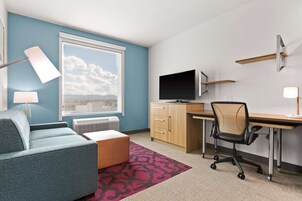 A modern room with a blue wall, a large window, a flat-screen TV, a desk, a chair, a sofa, and a patterned rug.