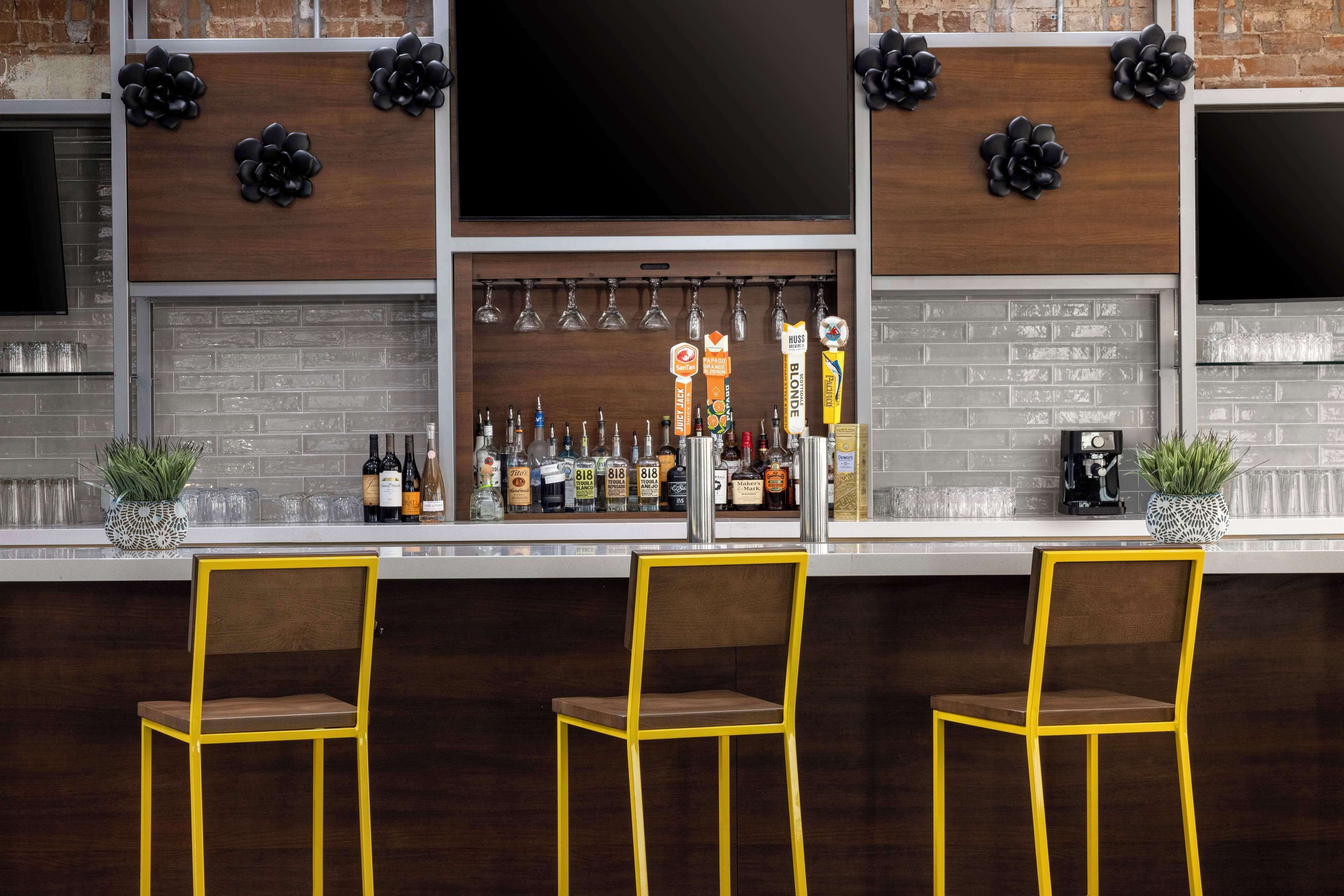 Uma área de bar com três cadeiras amarelas, uma seleção de garrafas e flores negras decorativas na parede.