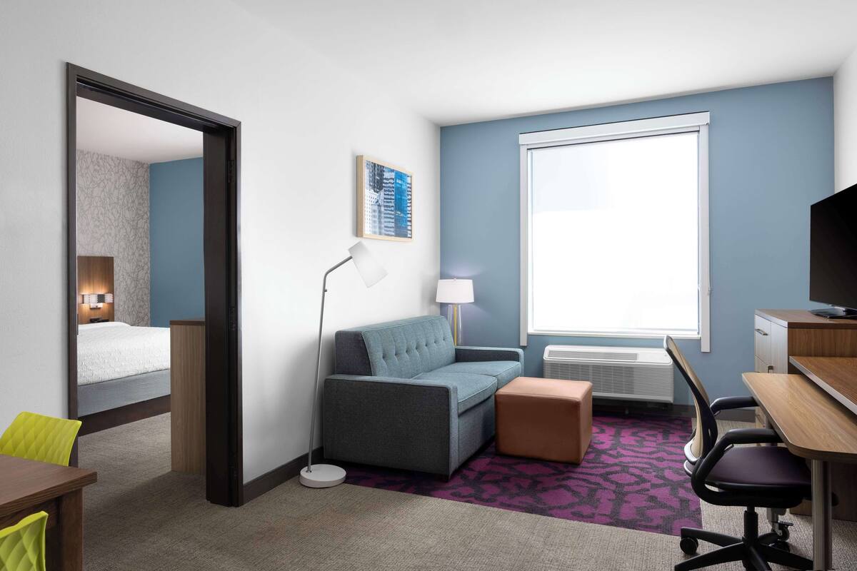 A hotel room with a bed, a sofa, a desk, a chair, a lamp, a TV, and a window.