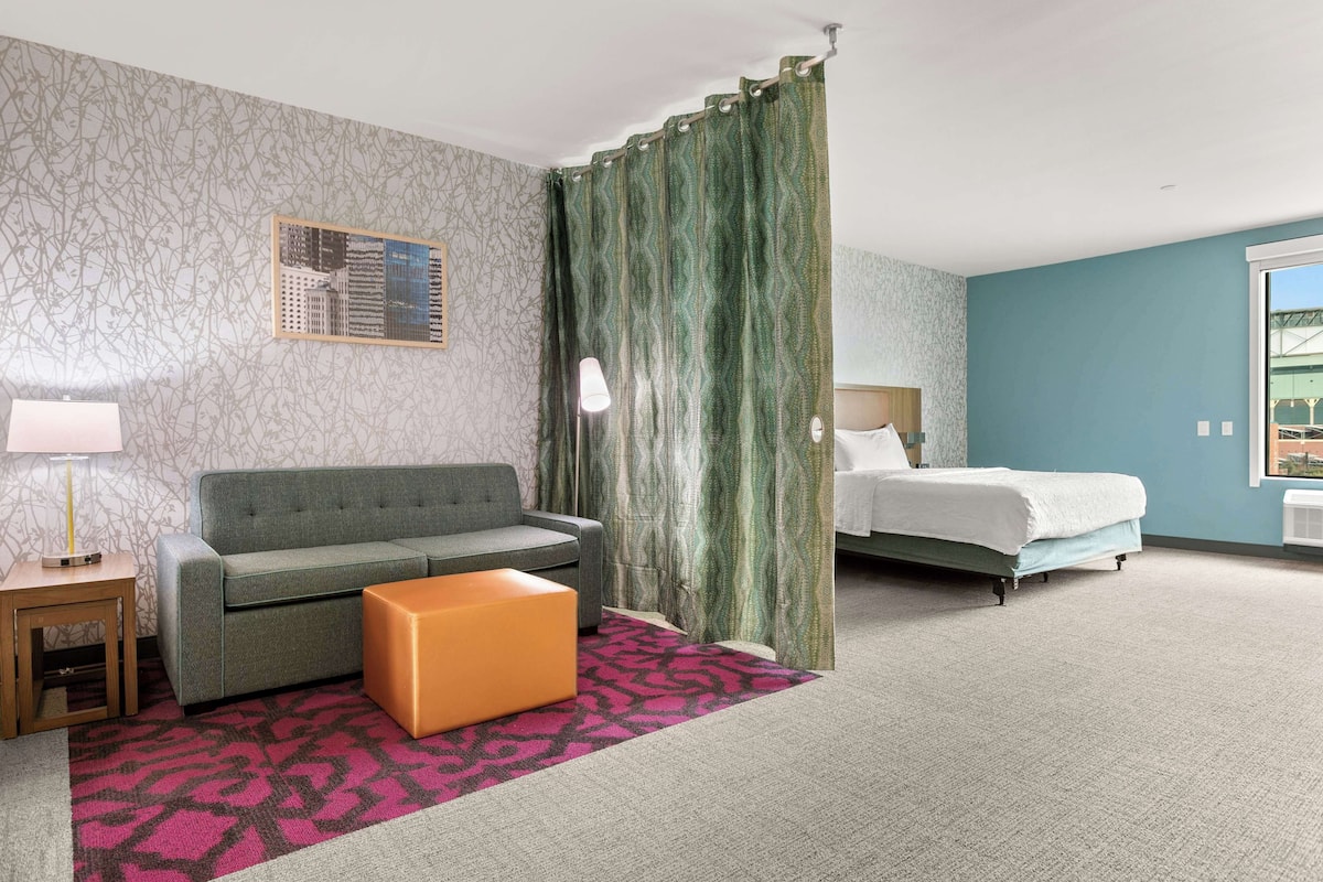 A hotel room with a grey sofa, an orange ottoman, a bed with white bedding, a green curtain, and a cityscape view.