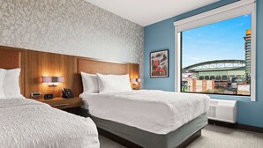 A hotel room with two beds, a large window with a city view, a wooden headboard, and a nightstand with a phone and lamp.