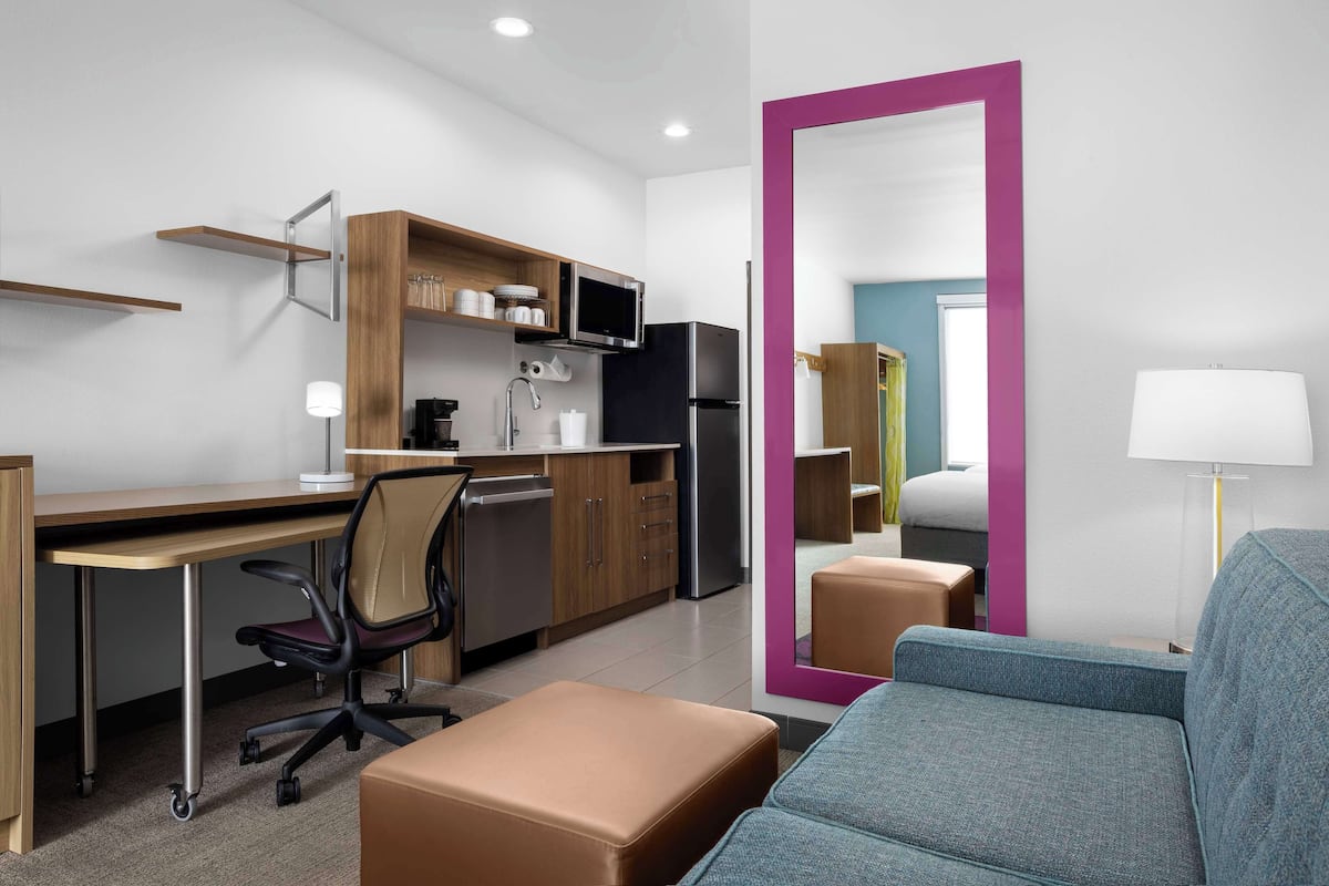 A modern hotel room with a kitchenette, a sofa, a desk, and a large mirror.